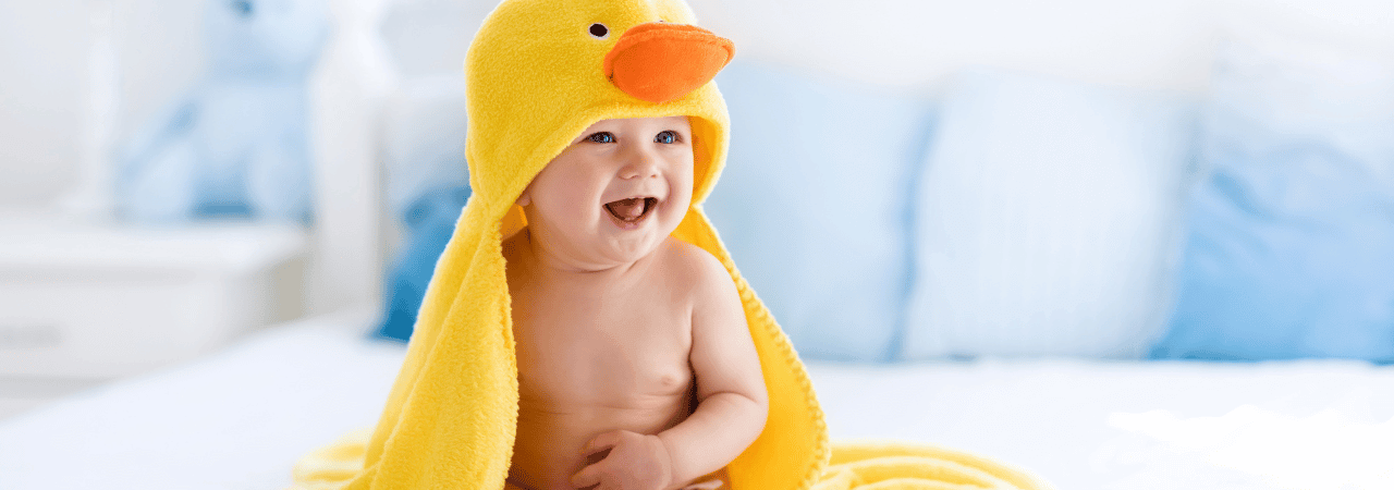 Smiling baby wearing a yellow duck hood towel on a bed